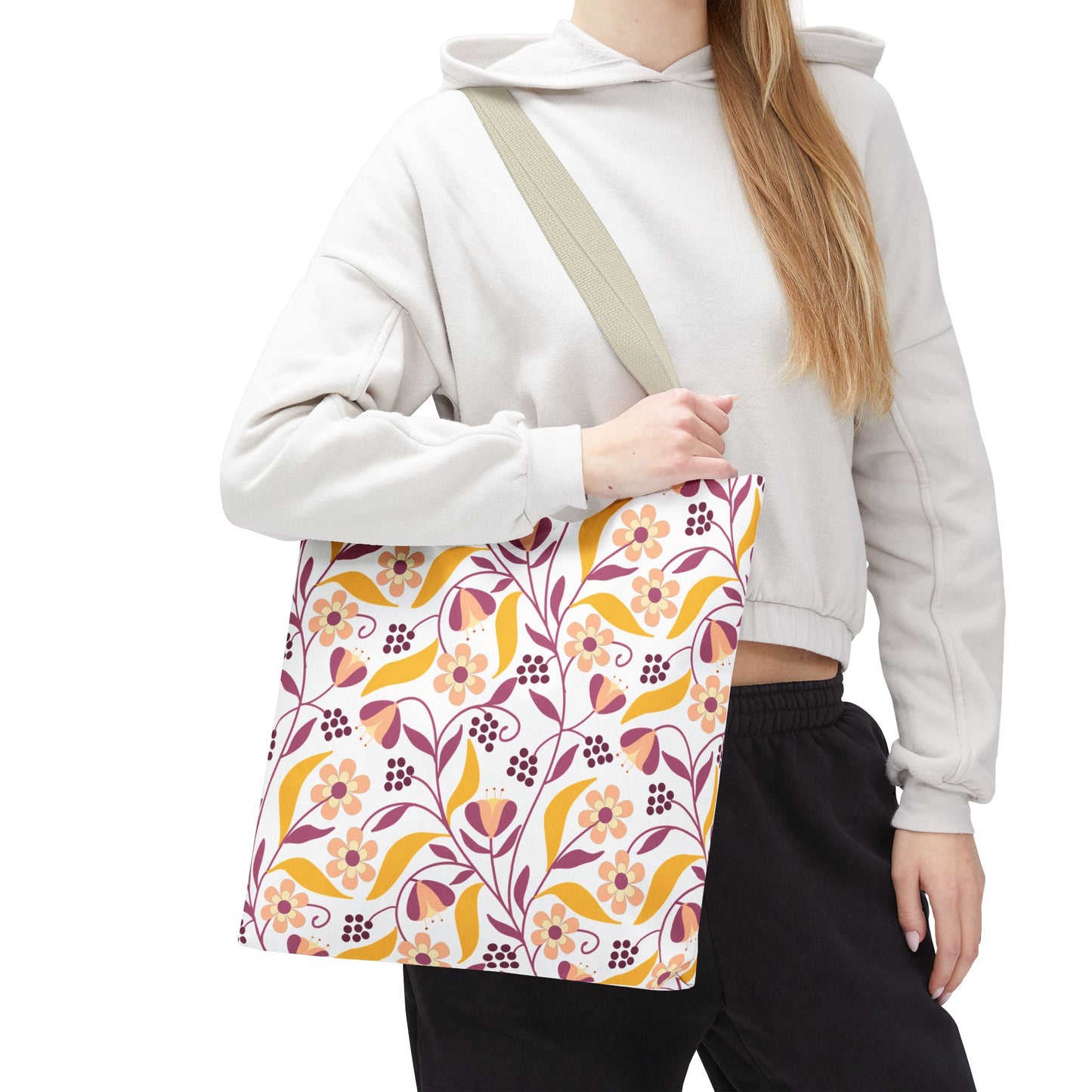 Person holding the Floral Tote Bag, featuring a white base with warm-toned floral patterns. It hangs from a light-beige strap, complementing the person's casual attire of a gray hoodie and black pants.