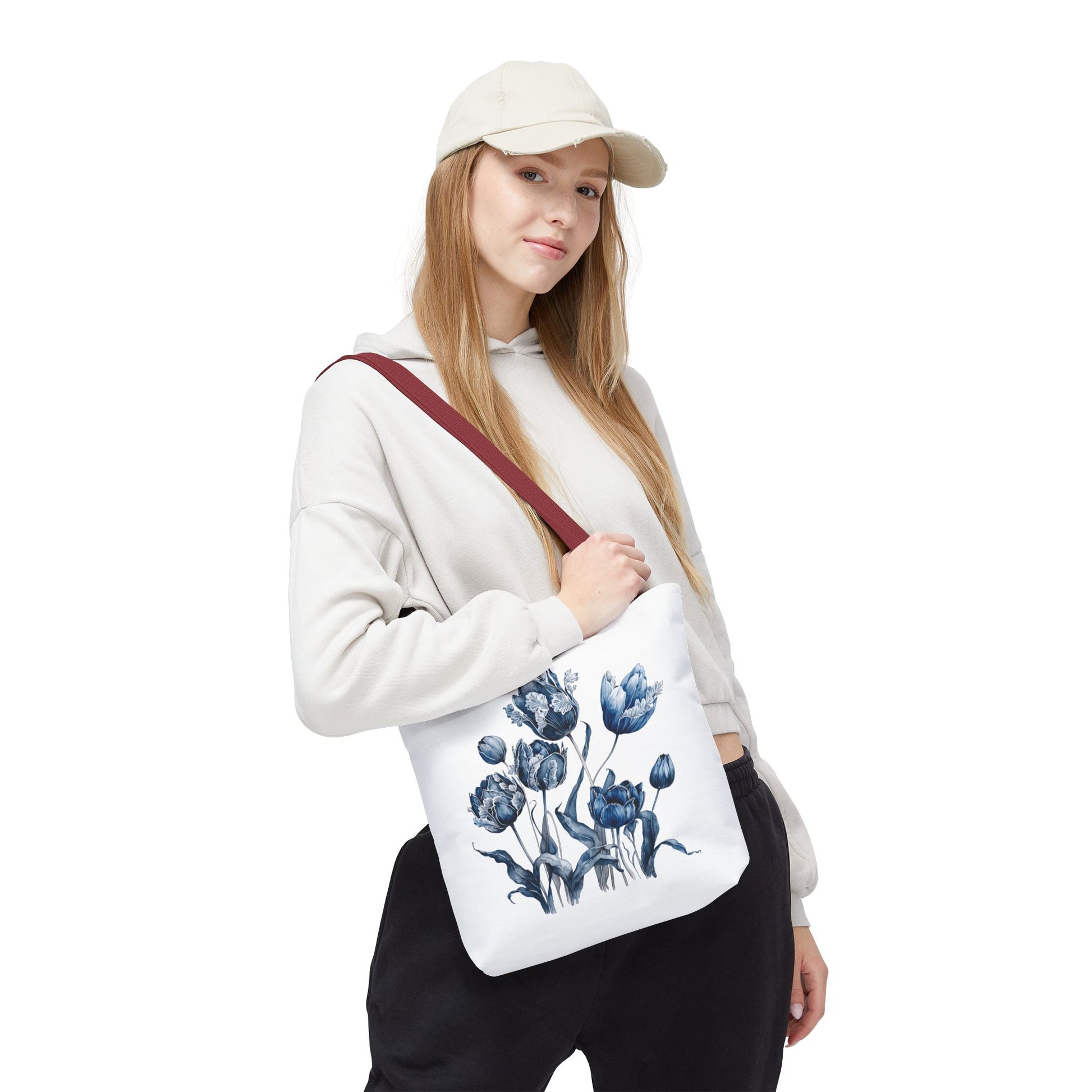 Woman wearing a hat and sweatshirt holds the Flowers in Blue Hue Watercolor Tote Bag, showcasing its floral design and sturdy construction.