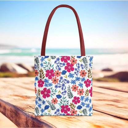 Confetti Wildflower Tote Bag with a vibrant watercolor floral print on a white background, featuring reddish-brown straps, displayed on a wooden surface with a soft-focus beach backdrop. Ideal for joyful summer adventures.