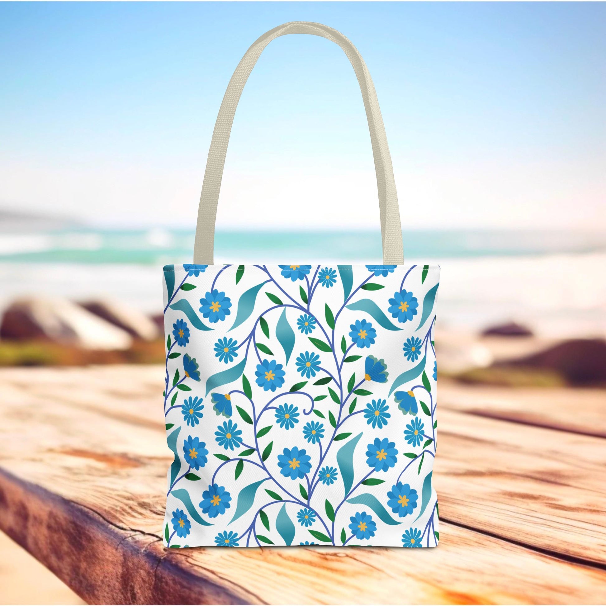 Blue Floral Tote Bag on a wooden boardwalk, featuring a vibrant daisy pattern with blue flowers and green leaves. Perfect for summer outings, offering durability and style with reinforced handles and ample space.