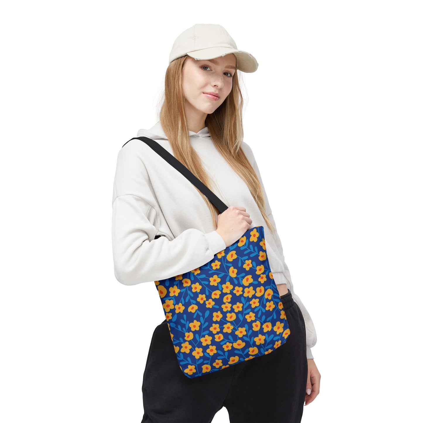 Cobalt Poppy Field Tote Bag featuring a vibrant pattern of yellow flowers with orange centers and blue leaves, carried casually over the shoulder by a young woman in a light hoodie and cap.