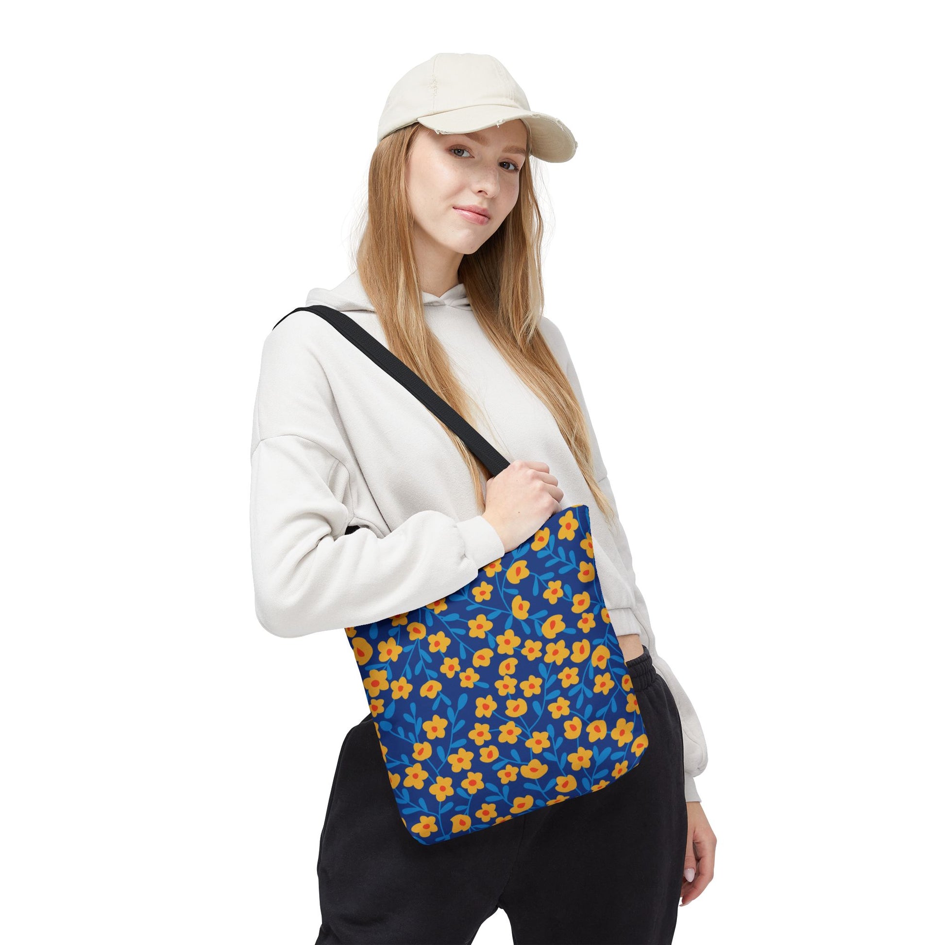 Cobalt Poppy Field Tote Bag featuring a vibrant pattern of yellow flowers with orange centers and blue leaves, carried casually over the shoulder by a young woman in a light hoodie and cap.