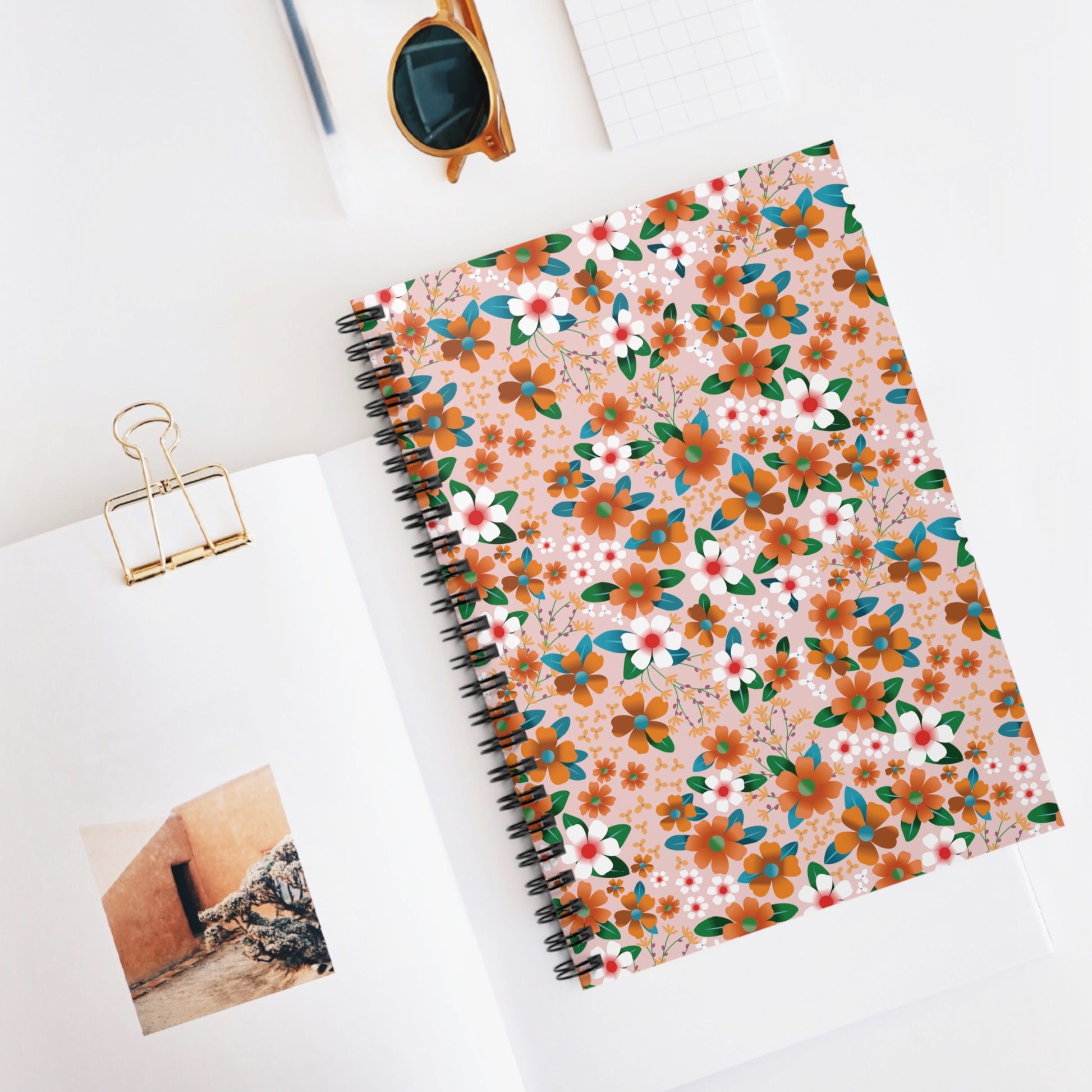 Floral Spiral Notebook with vibrant floral cover, centered on a desk setup with sunglasses and graph paper, embodies FireBrook Designs' blend of style and functionality for joyful note-taking.