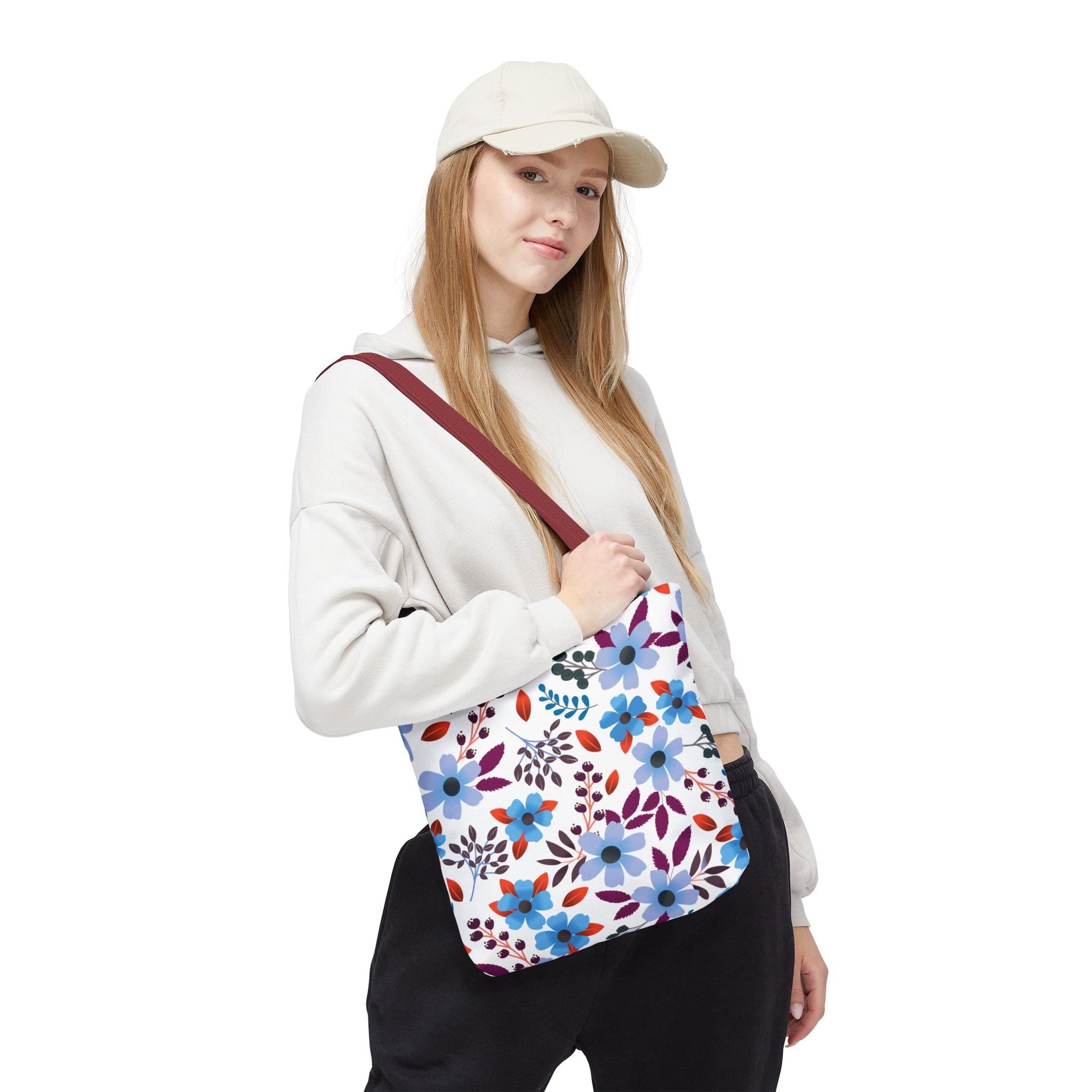 Floral Tote Bag held by a person wearing a light hoodie and cap, featuring a white base with blue, purple, and red blossoms, maroon strap, ideal for joyful summer outings.