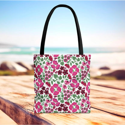 Pink Lady Tote Bag with black straps, showcasing a vibrant floral pattern of pink and burgundy blooms on white. Positioned on a wooden surface, it exudes a summery, beachside vibe.
