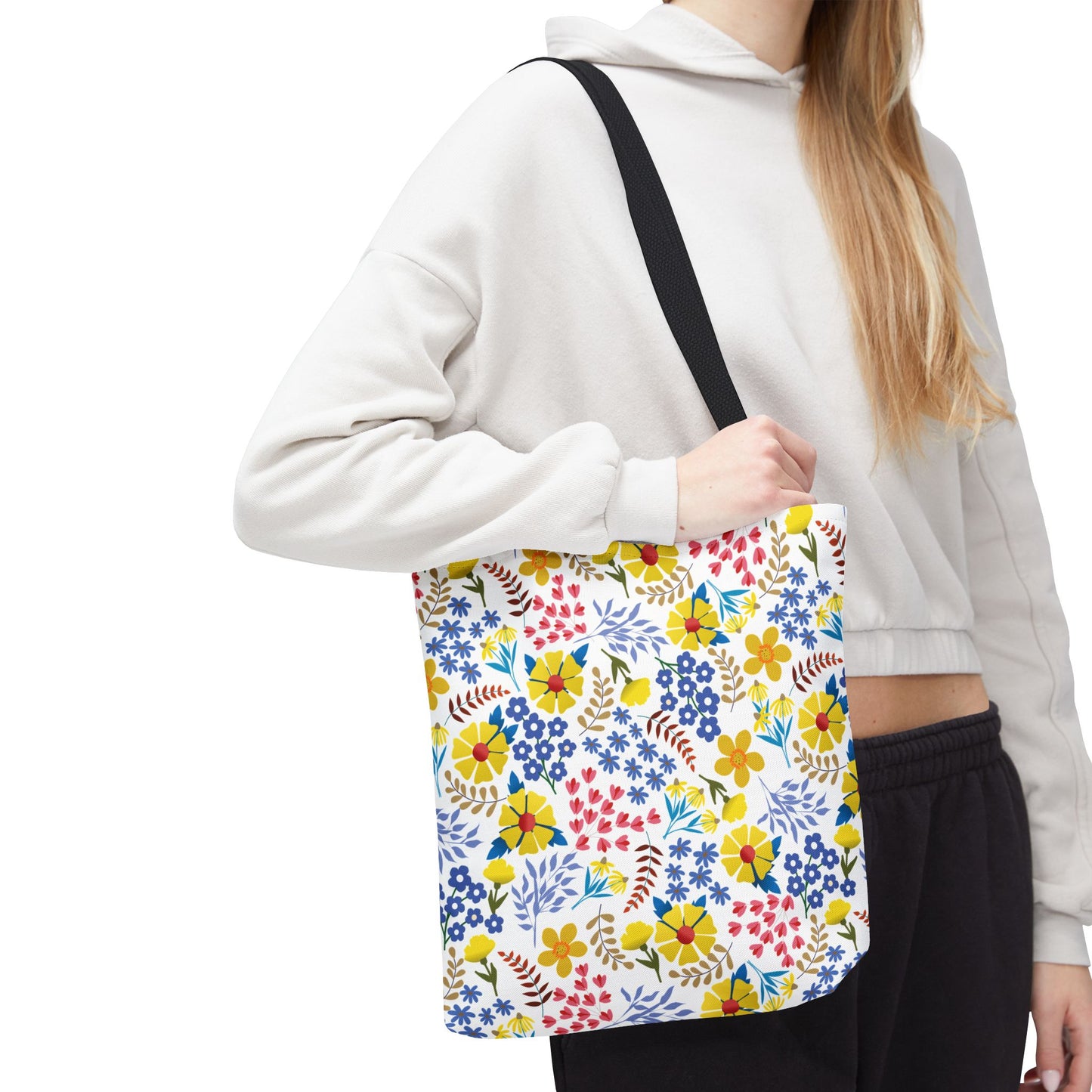 Person wearing a light cropped hoodie and dark pants holds a Floral Tote Bag with black straps and a vibrant floral pattern, embodying FireBrook Designs' cheerful and positive style for joyful summer outings.
