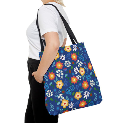 Person wearing a white T-shirt and black pants, carrying the Cornflower Patch Tote Bag. The bag features a vibrant floral pattern with red, yellow, and blue blossoms, reflecting FireBrook Designs' joyful style.