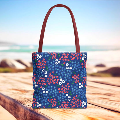 Berry Blue Festival Tote Bag featuring a vibrant floral pattern with red, white, and blue blossoms, set on a wooden surface with beach backdrop; ideal for joyful summer outings.