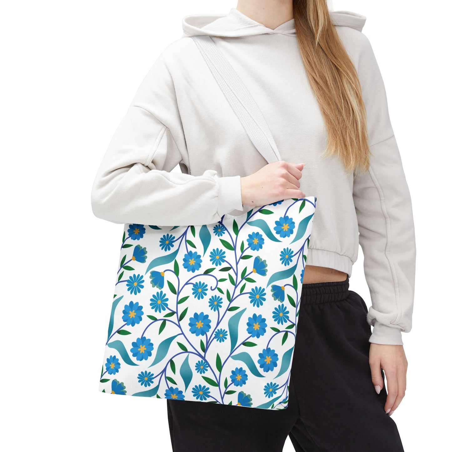 Blue Floral Tote Bag held by a person in a light-gray hoodie and black pants, featuring bright blue flowers with yellow centers, green leaves, and teal ribbons on a white canvas, perfect for joyful outings.