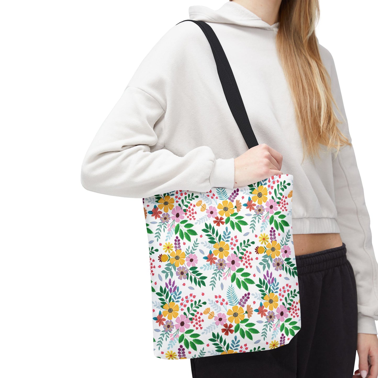 Person holding the Floral Tote Bag with bright floral pattern and black strap, wearing a cream hoodie and dark sweatpants. Ideal for vibrant summer outings, reflecting FireBrook Designs' joyful and uplifting aesthetic.