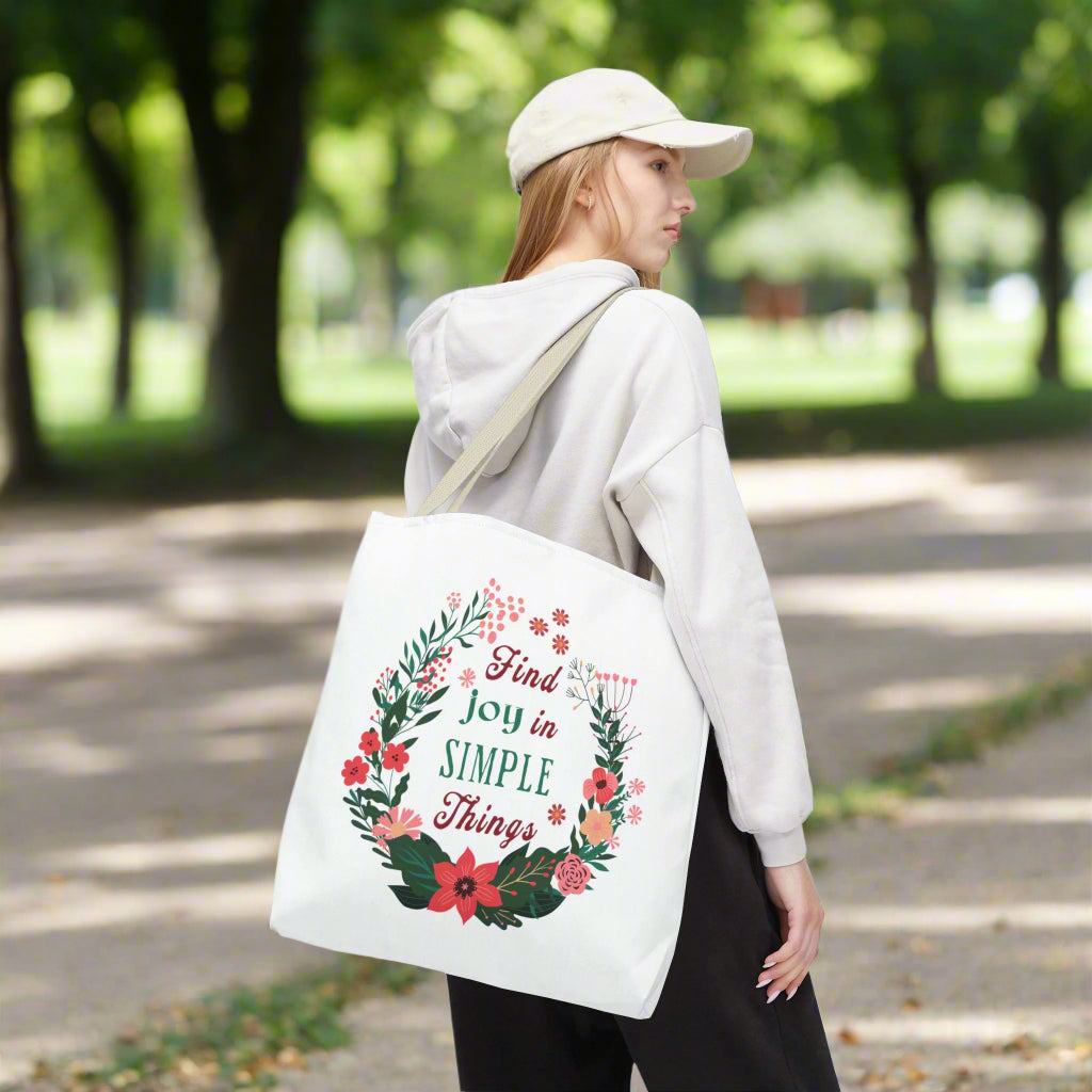 Woman wearing a white hat and sweatshirt, holding the Find Joy in Simple Things Tote Bag featuring a watercolor floral design, ideal for daily use.
