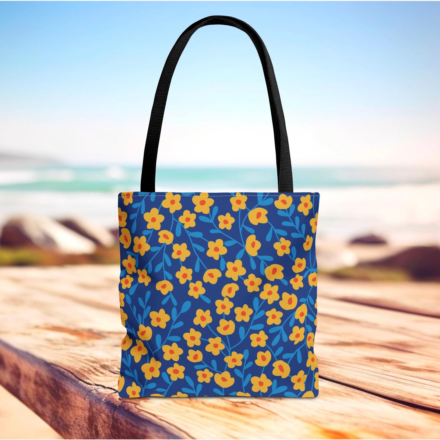 Floral Tote Bag with black handles, featuring yellow-orange flowers and blue leaves pattern on a deep blue background, positioned on a wooden surface, perfect for brightening summer outings and beach trips.