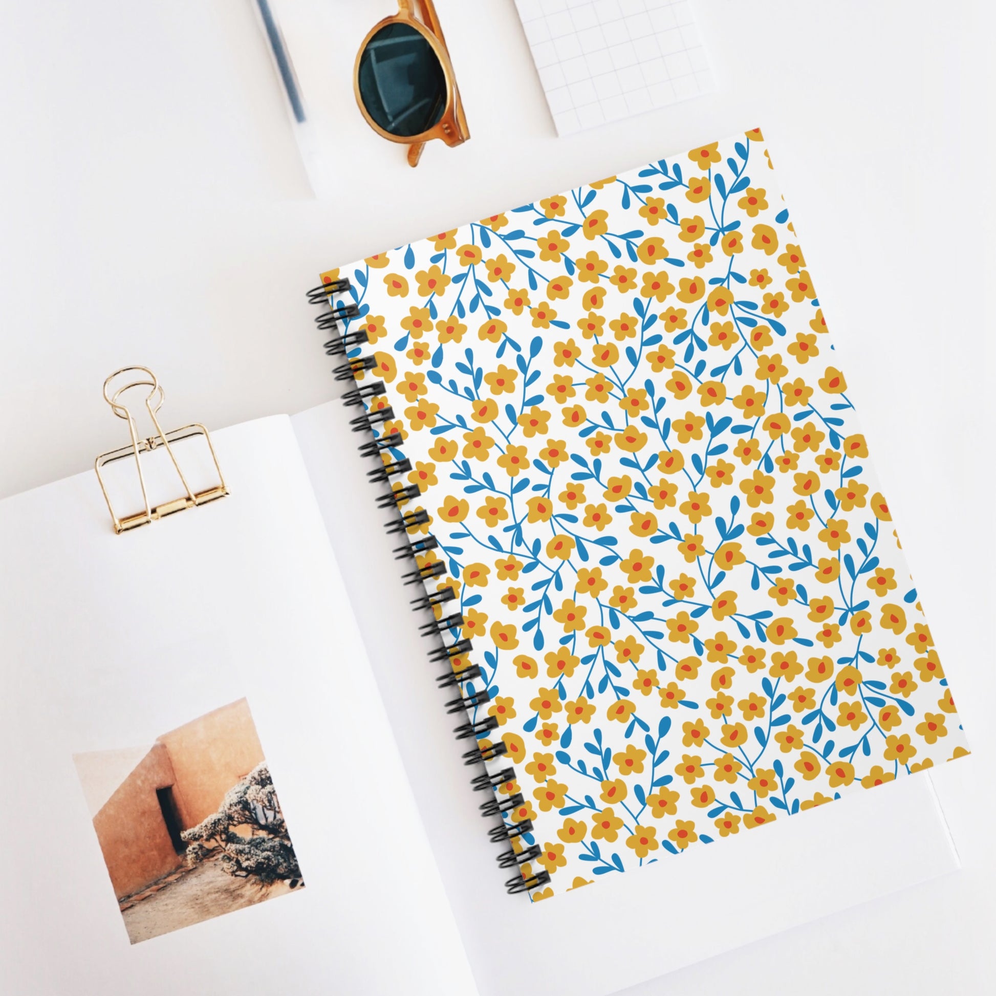 Floral Spiral Notebook with yellow flowers and blue stems on the cover, surrounded by an open magazine, sunglasses, and graph paper, embodying the joyful essence of FireBrook Essentials.