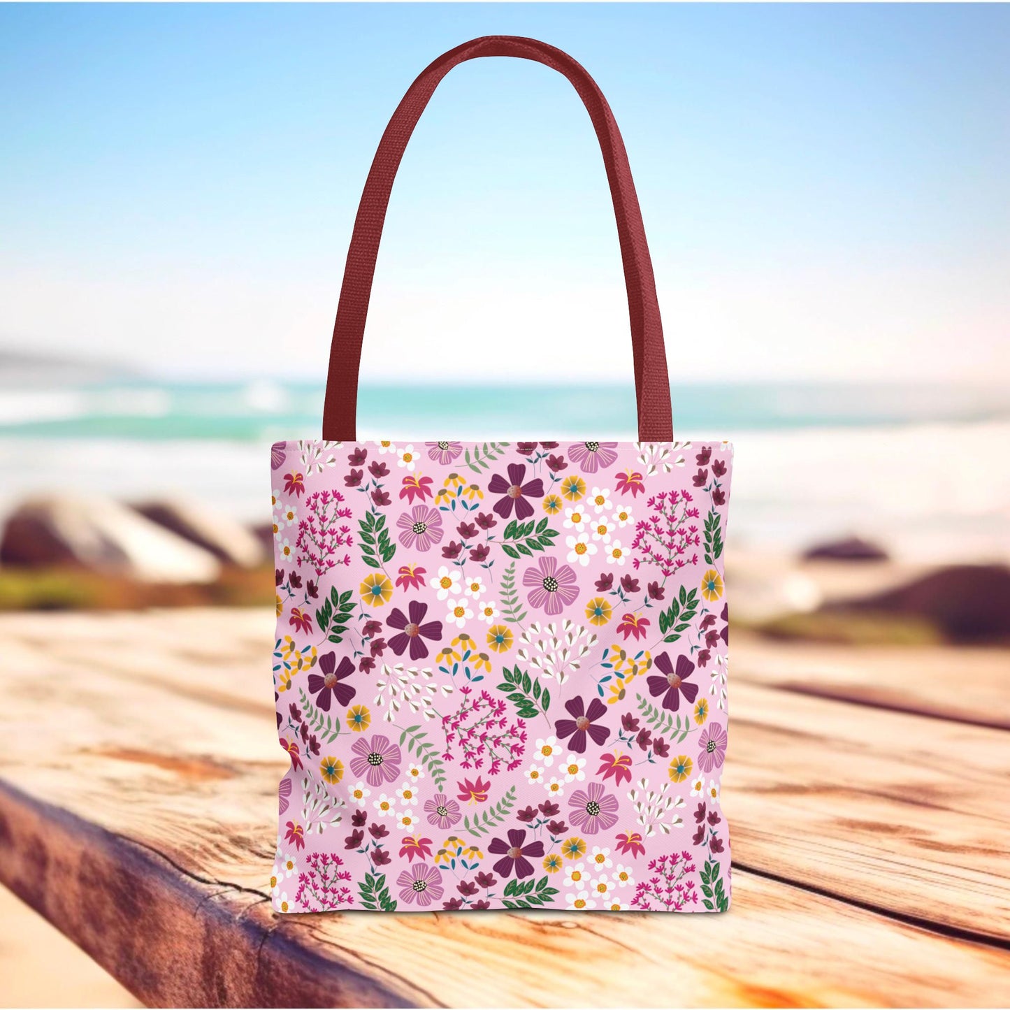 Floral Tote Bag with a pink base and vibrant blossoms, featuring burgundy handles, rests on a wooden surface. Ideal for daily essentials, it embodies FireBrook Designs' commitment to stylish, durable solutions.