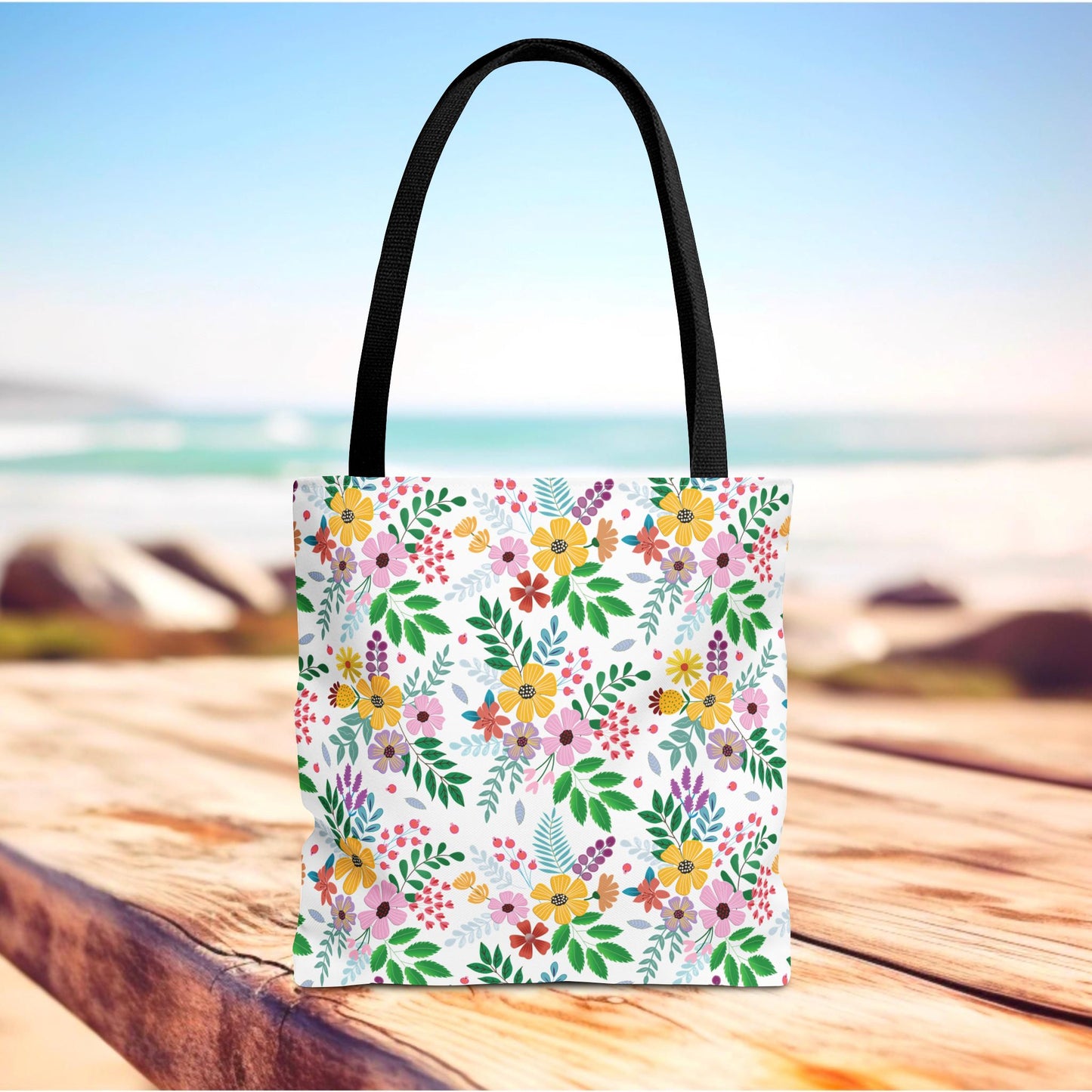 Floral Tote Bag with colorful flower pattern and black straps, resting on a wooden surface, evoking a summery, seaside vibe. Perfect for vibrant, joyful outings from FireBrook Designs.