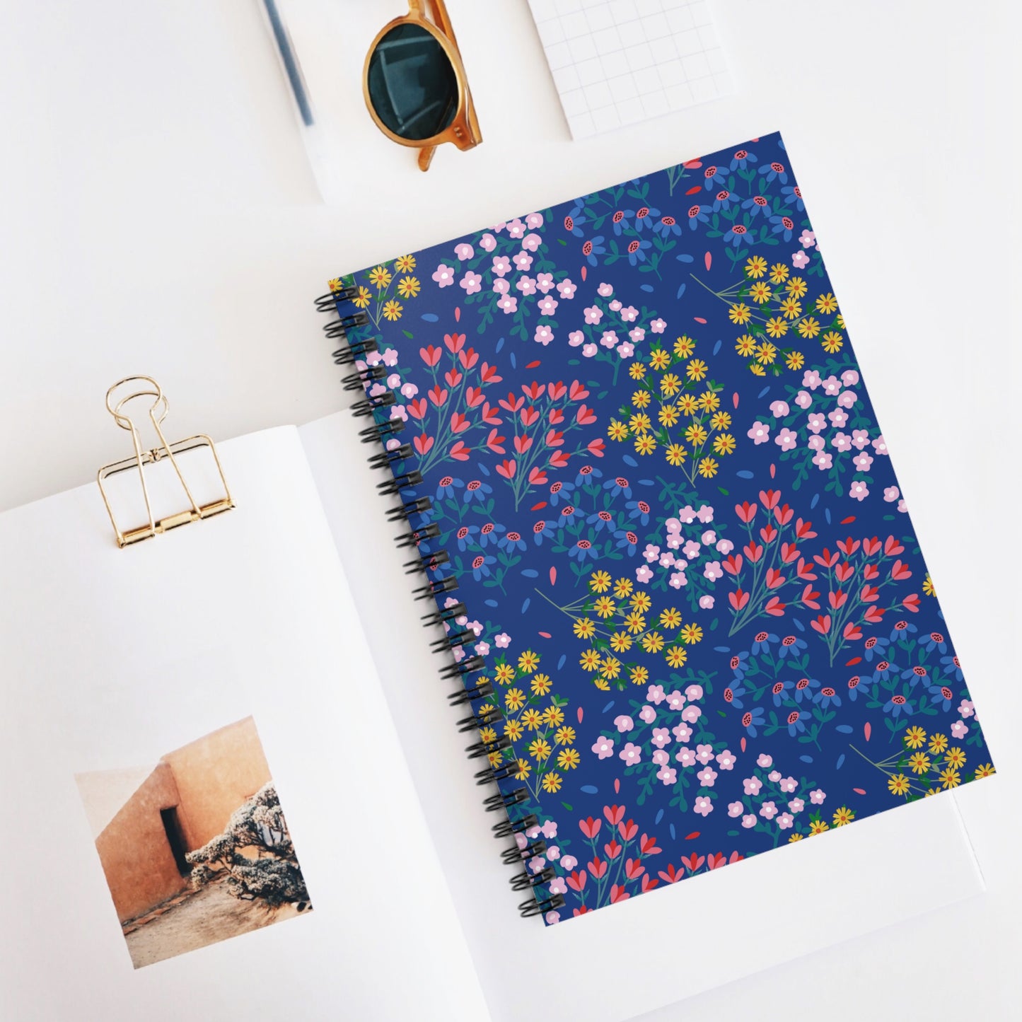 Floral Spiral Notebook with dark blue cover featuring colorful floral motifs, positioned on a desk with an open magazine and sunglasses, designed for creative expression and organization. Ideal for students and professionals.