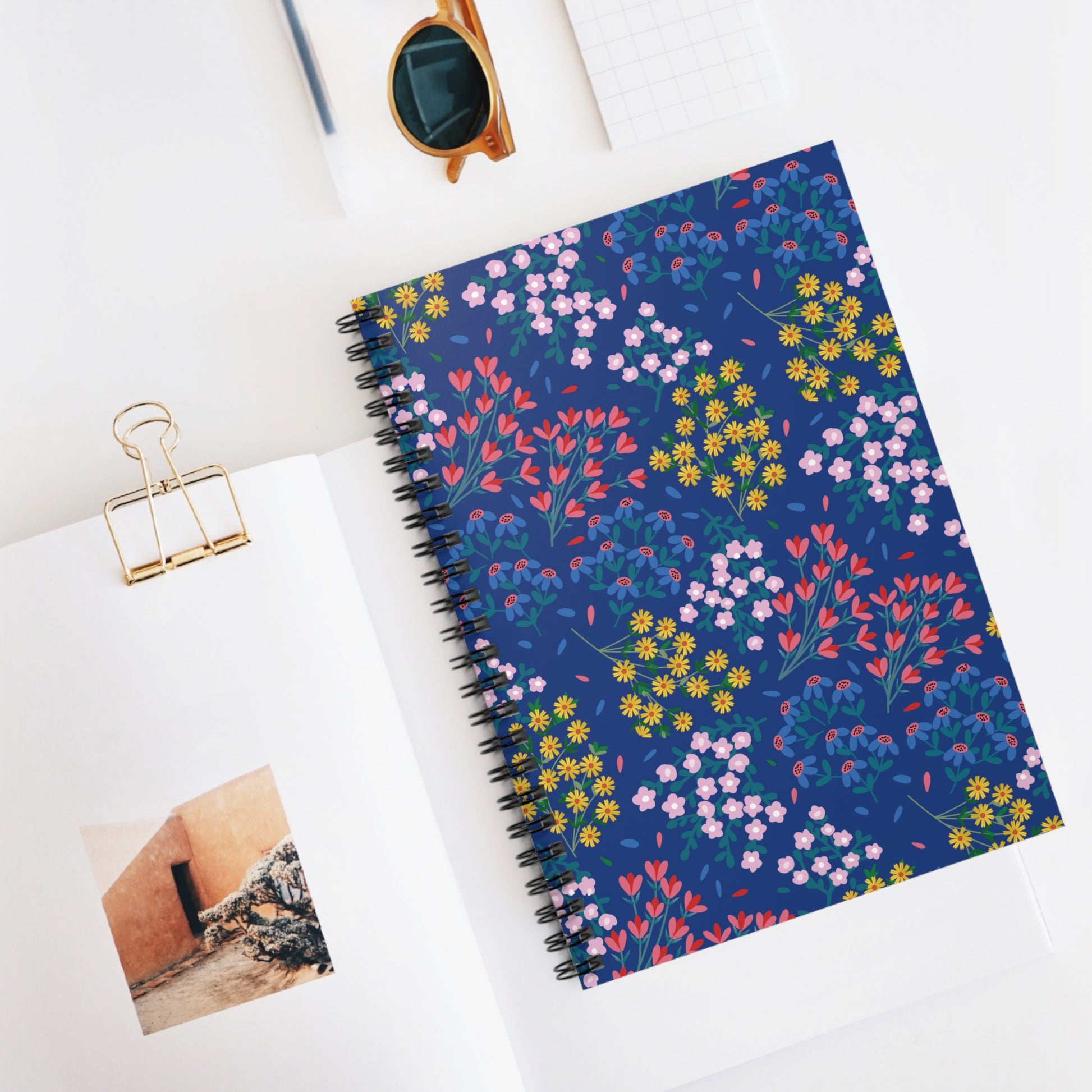 Floral Spiral Notebook with dark blue cover featuring colorful floral motifs, positioned on a desk with an open magazine and sunglasses, designed for creative expression and organization. Ideal for students and professionals.