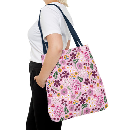 The Rose Garden Blush Tote Bag, featuring a floral pattern with purple, white, yellow, and pink blossoms, is carried by a person. It has navy-blue straps and complements their casual outfit.