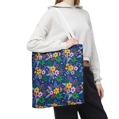 Person wearing a light hoodie and dark pants holds a Floral Tote Bag with vibrant flowers and green leaves. The navy-blue bag, ideal for bright summer outings, hangs from a white shoulder strap.