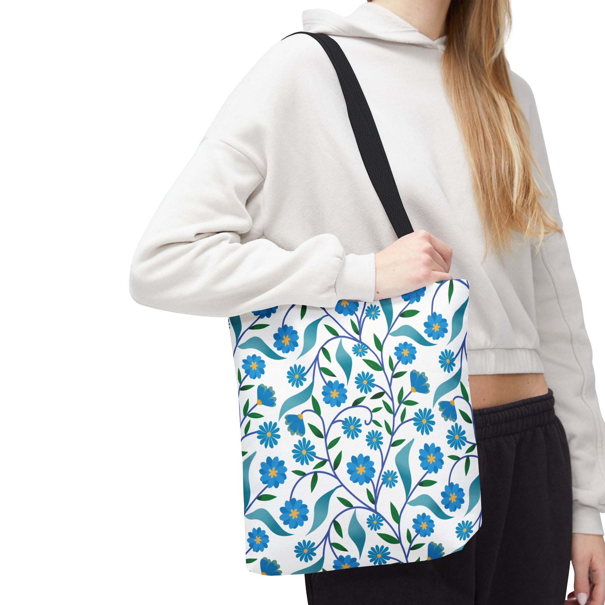 Person wearing a light hoodie and dark pants carries a Blue Floral Tote Bag featuring stylized blue flowers and green leaves with a black strap, embodying FireBrook Designs' cheerful, uplifting style.