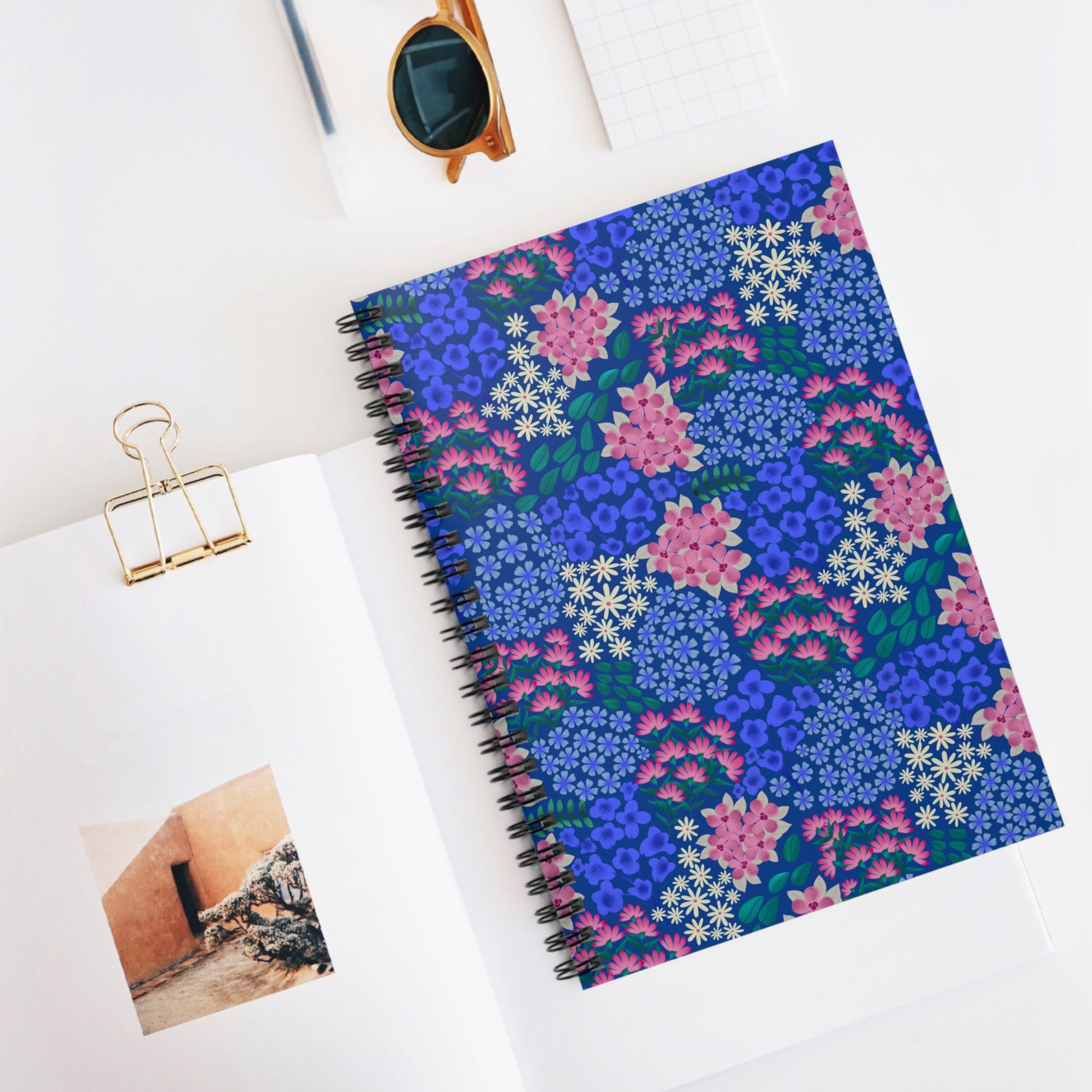 Floral Spiral Notebook with vibrant blue floral cover, surrounded by a book page with binder clip, photo, and sunglasses; perfect for sparking creativity and organizing thoughts.