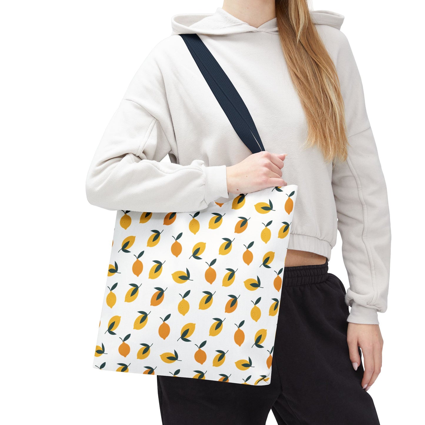Person holding the Lemon Drop Toss Tote Bag, showcasing a vibrant lemon design with green leaves on a white background, perfect for stylish, cheerful outings. Worn with a light hoodie and dark pants.