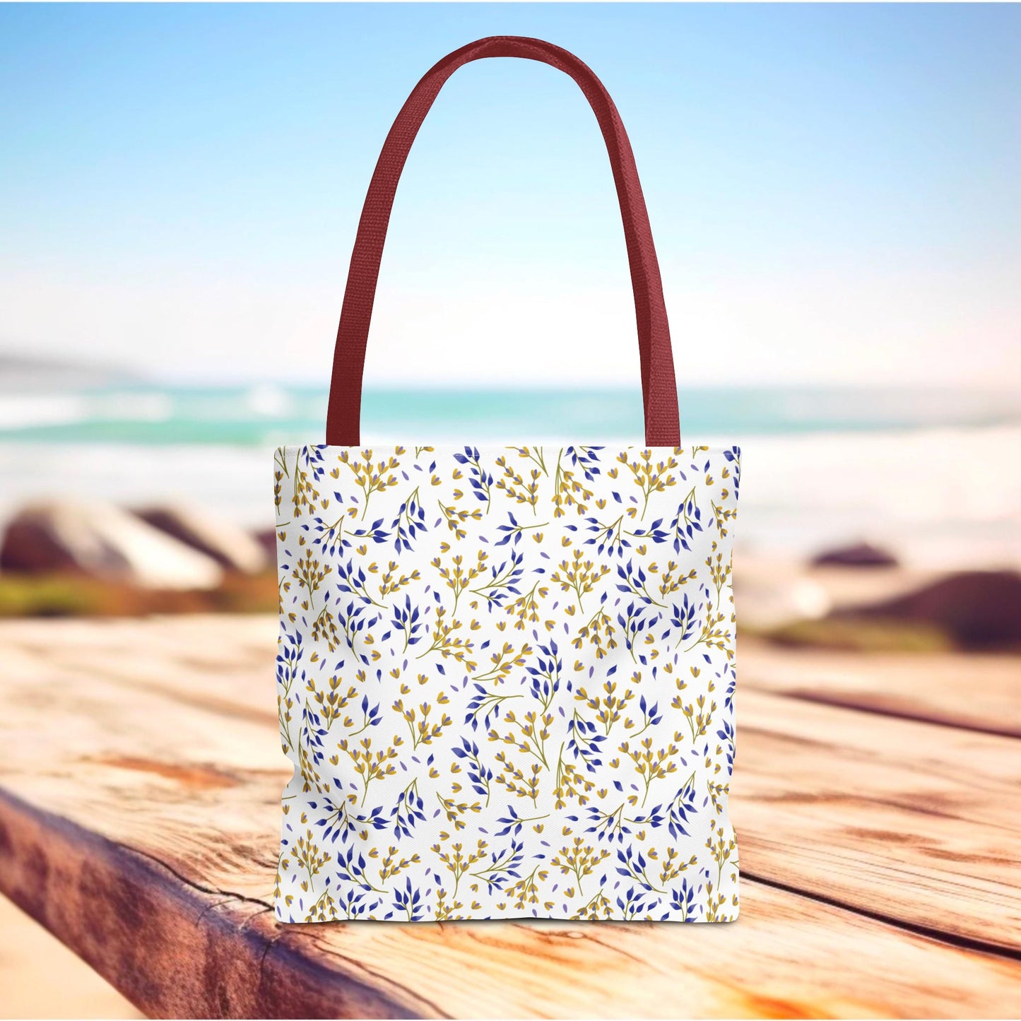 Wild Herb Garden Tote Bag with burgundy-red handles and a white body, adorned with gold and deep blue botanical patterns, standing on a boardwalk by the seaside, ideal for summer outings.