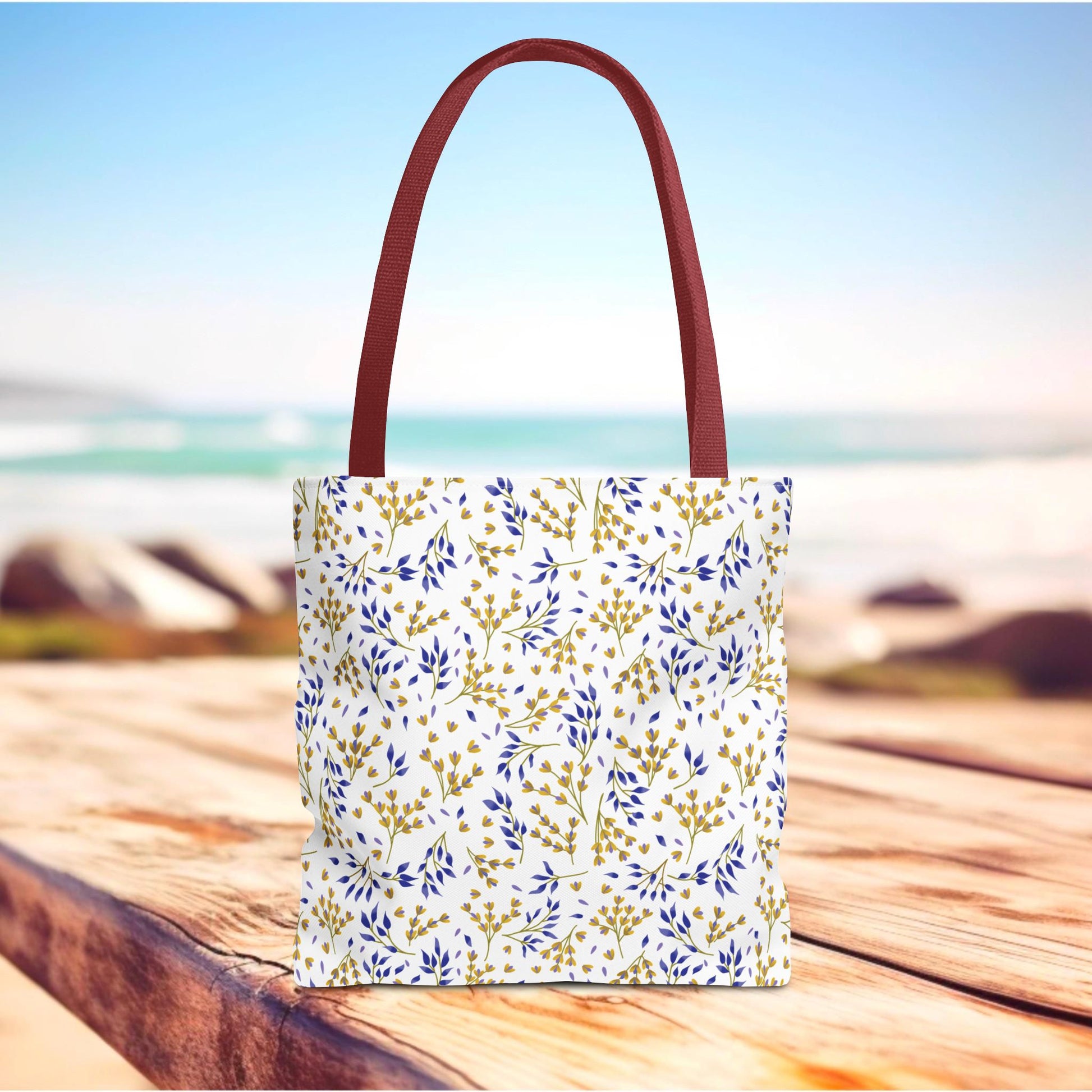 Wild Herb Garden Tote Bag with burgundy-red handles and a white body, adorned with gold and deep blue botanical patterns, standing on a boardwalk by the seaside, ideal for summer outings.