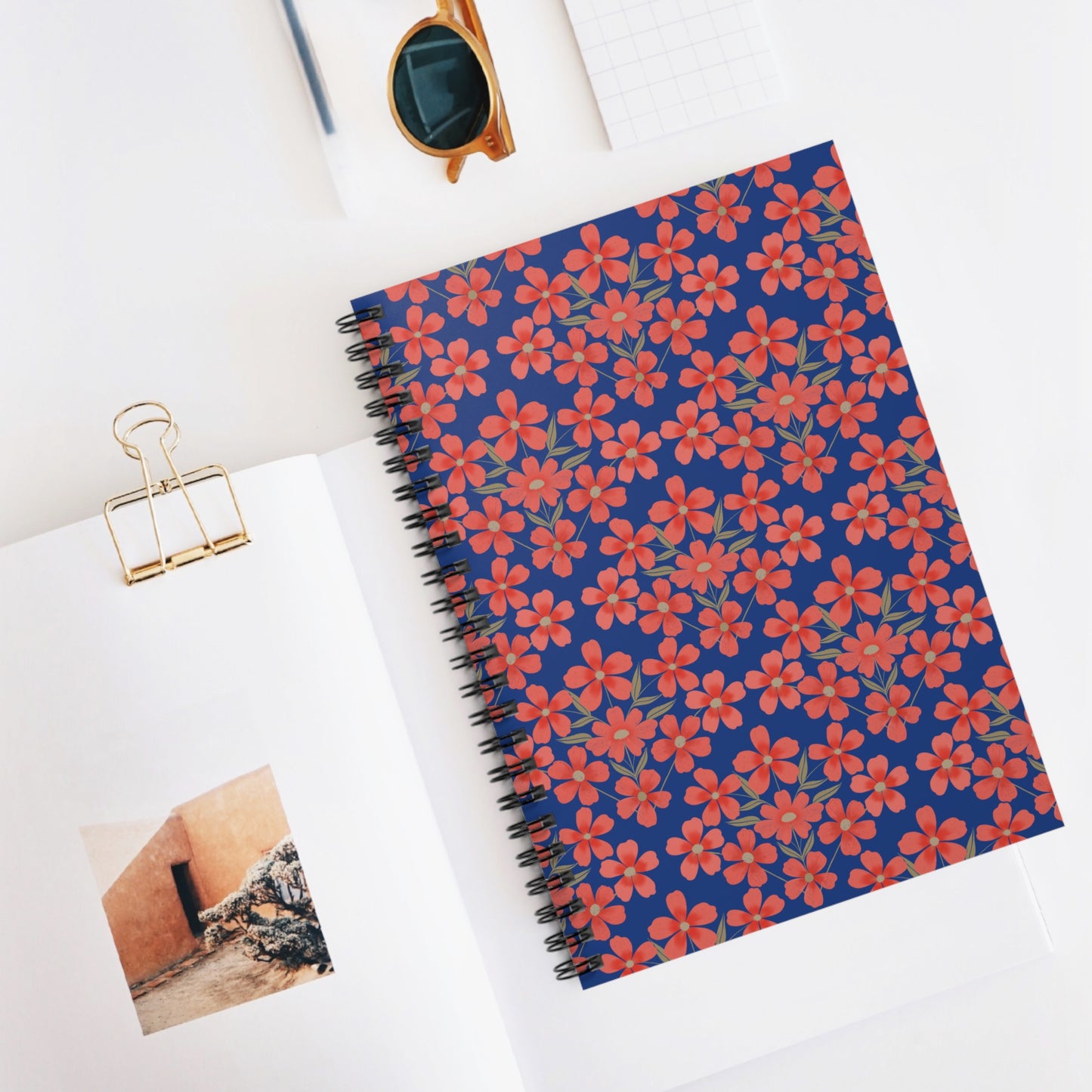 Floral Spiral Notebook with red-orange flowers and greenish leaves on a deep blue cover, surrounded by a magazine, gold clip, grid notepad, and brown sunglasses, embodying FireBrook Designs' joyful style.