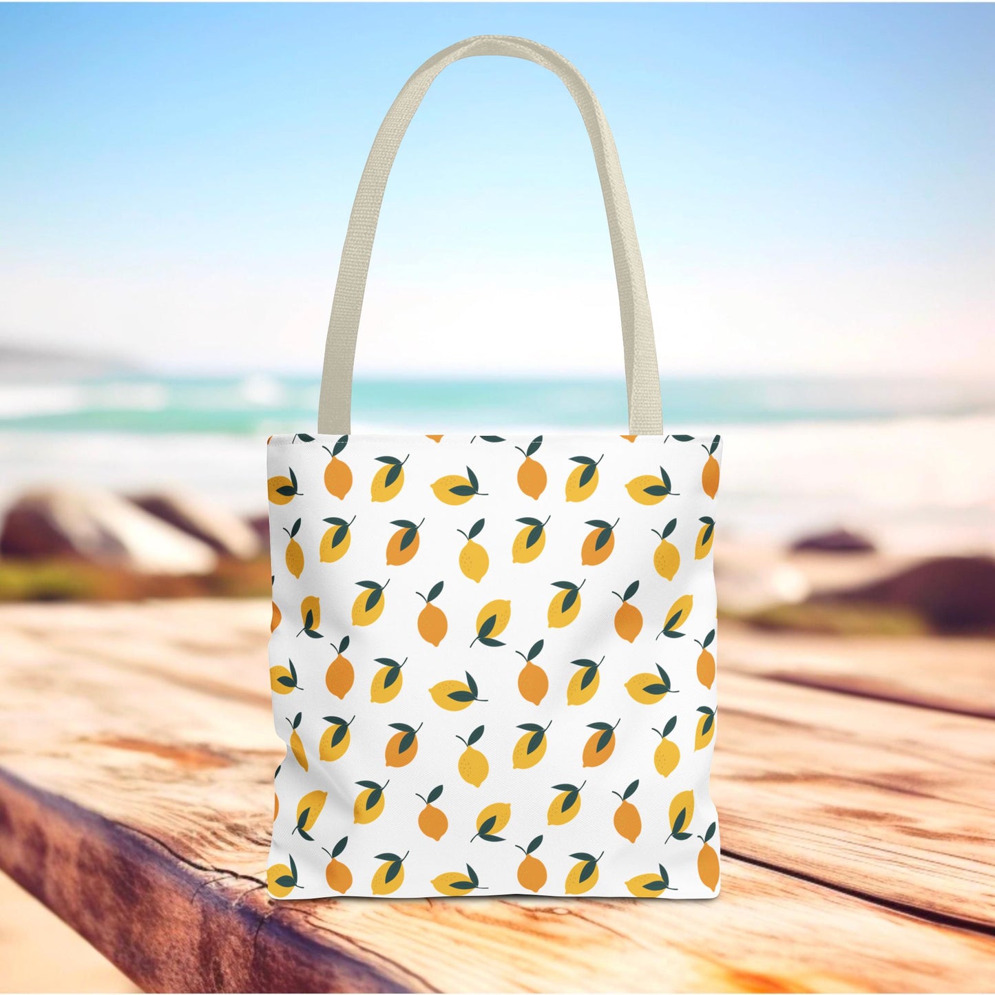 Lemon Drop Toss Tote Bag featuring vibrant yellow lemons and green leaves on white canvas, perfect for summer outings. Stands on a weathered wooden surface with a shoreline and ocean in the background.