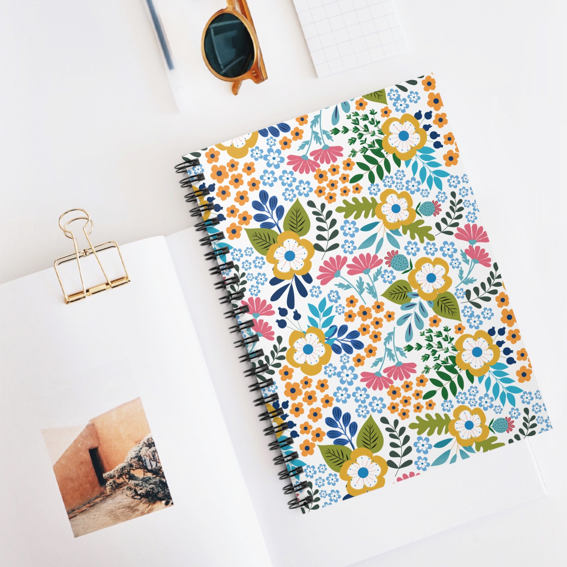 Floral Spiral Notebook with vibrant floral cover, central on a bright workspace flat lay, accompanied by round sunglasses, a grid notepad, open magazine, and pen, embodying FireBrook Designs' joyful style.