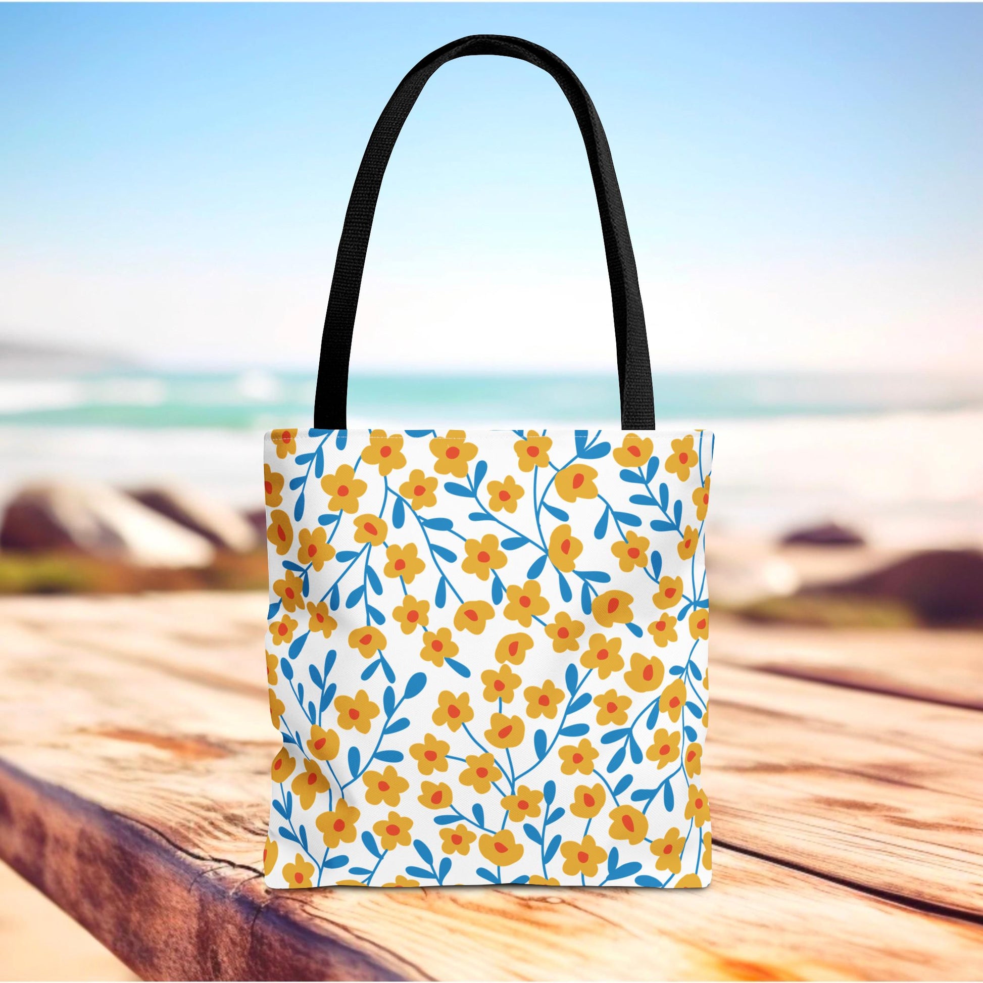 Floral Tote Bag on wooden surface with black straps, featuring yellow-orange flowers and blue stems, ideal for summer outings, embodying FireBrook Designs' uplifting style and joyful, practical design.