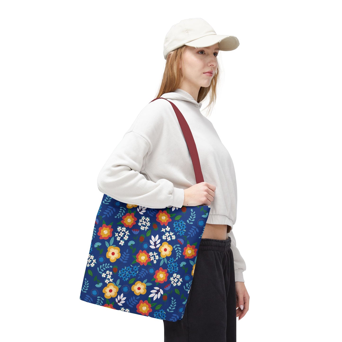 Person with a Cornflower Patch Tote Bag featuring a vibrant floral pattern and reddish strap, perfect for joyful summer outings. They wear a light-gray hoodie and dark pants, embodying FireBrook Essentials' uplifting style.