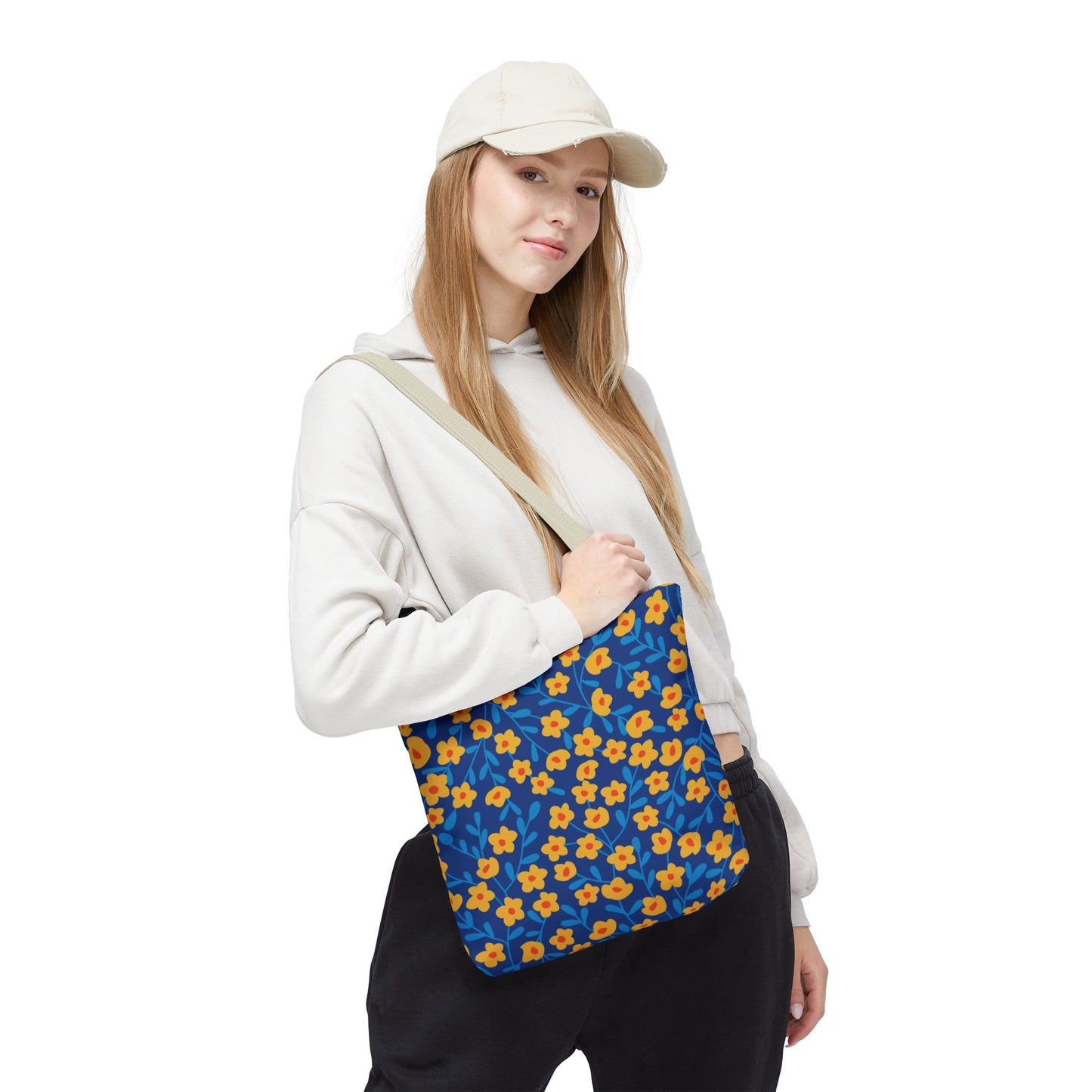 Young woman in a light hoodie carries a Cobalt Poppy Field Tote Bag, showcasing a vibrant blue background with yellow-orange flowers. Perfect for summer outings, reflecting FireBrook Designs' uplifting, joyful style.