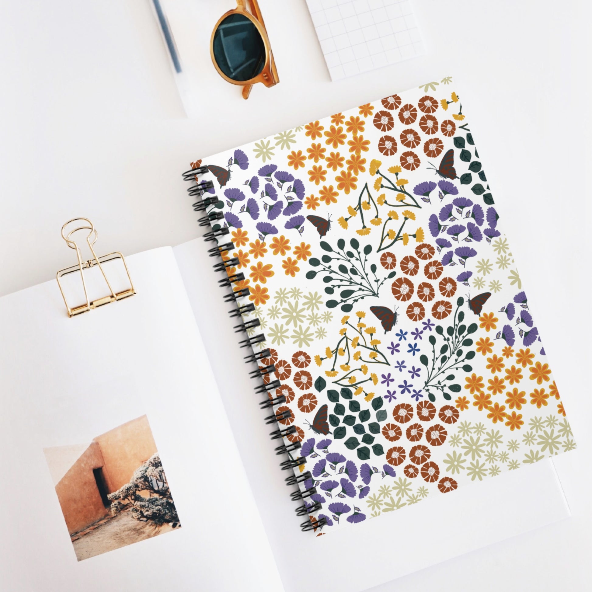 Floral Spiral Notebook with vibrant flower and butterfly design on a white cover, atop an open book. Ideal for creative journaling, note-taking, or sketching, embodying FireBrook Designs' uplifting style.