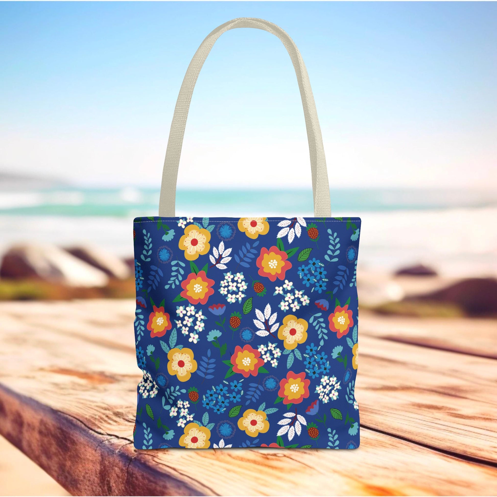 Floral Tote Bag with a bold blue body featuring vibrant yellow, red, and white blossoms, sits on a wooden surface by the seaside. Ideal for joyful summer outings, reflecting FireBrook Designs' vibrant style.