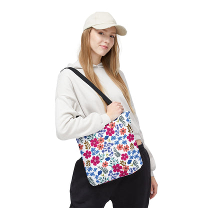 A young woman models the Floral Tote Bag, showcasing its vibrant pink, blue, and orange floral pattern with green leaves. The bag's black strap contrasts her light hoodie and complements her casual style.