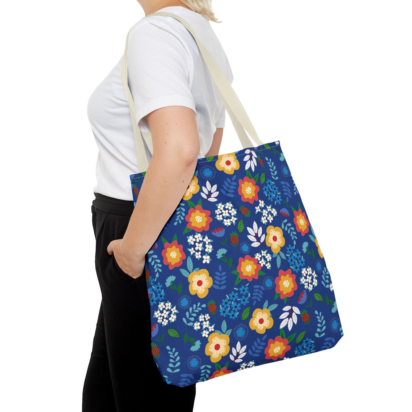 Person wearing a white T-shirt and black pants carries the Cornflower Patch Tote Bag, featuring a bright blue background with a vibrant floral pattern, ideal for cheerful and stylish summer outings.