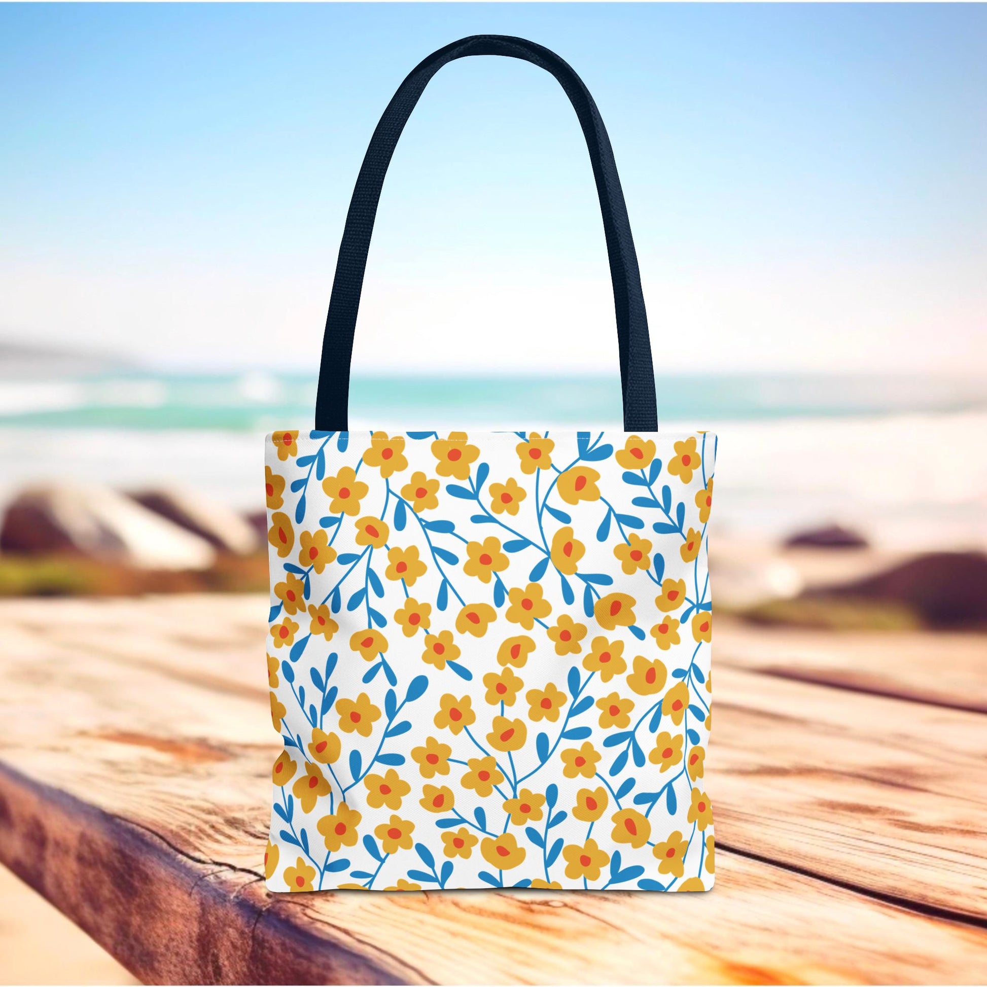 Floral Tote Bag with vibrant yellow-orange blossoms and blue stems on a white base, set against a beach backdrop, ideal for joyful summer outings. Features black handles and reinforced stitching for durability.