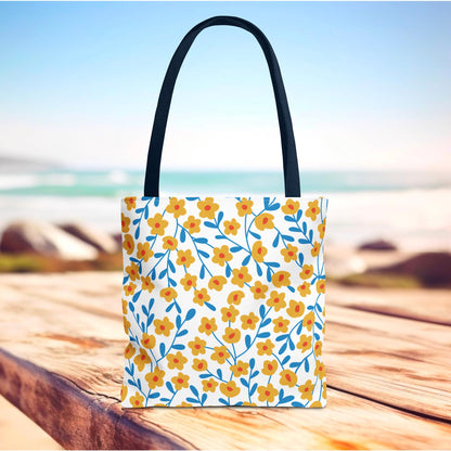 Floral Tote Bag with vibrant yellow-orange blossoms and blue stems on a white base, set against a beach backdrop, ideal for joyful summer outings. Features black handles and reinforced stitching for durability.