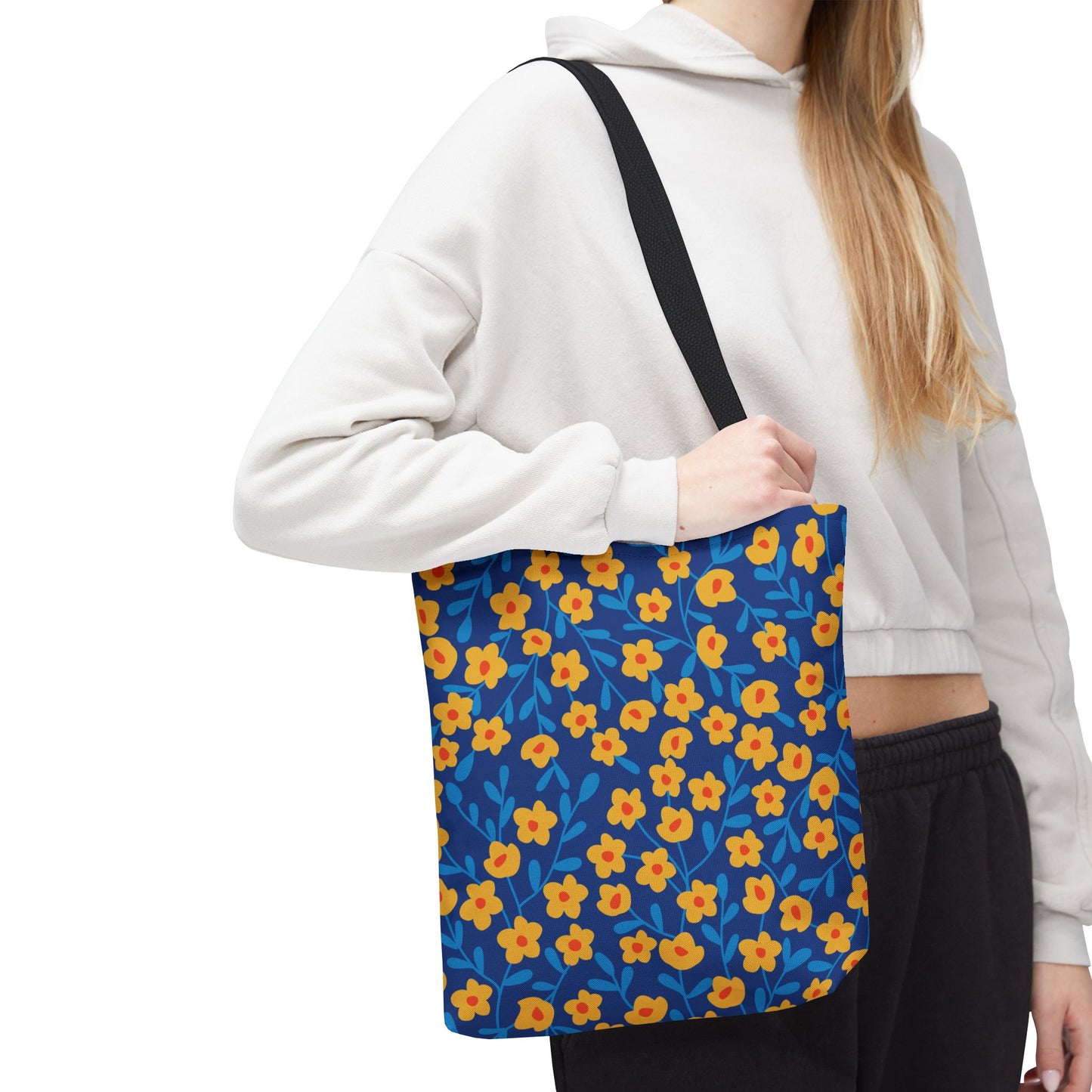 Person wearing a light hoodie and dark pants holds the Cobalt Poppy Field Tote Bag, adorned with yellow-orange flowers and blue leaves, perfect for joyful, stylish summer outings.