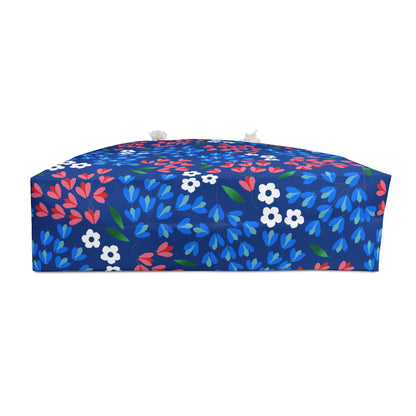 Weekender Bag: A low, rectangular pouf with a mid-blue fabric featuring stylized blossoms and leaves, complete with domed top, tassels, and box-shaped sides. Perfect for stylish, joyful travel.