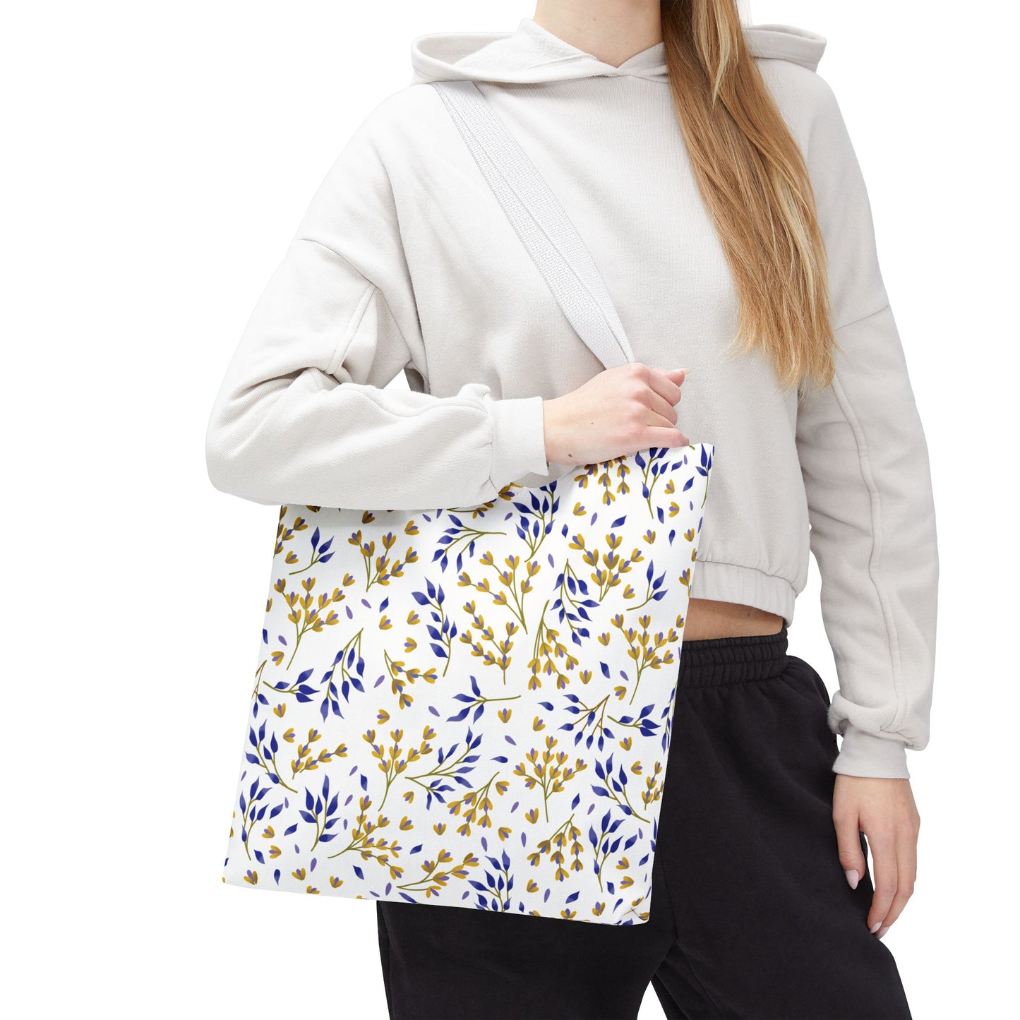Person wearing a light hoodie and dark pants, holding a Floral Tote Bag with a white background, yellow blossoms, and blue leaves pattern, ideal for summer outings.