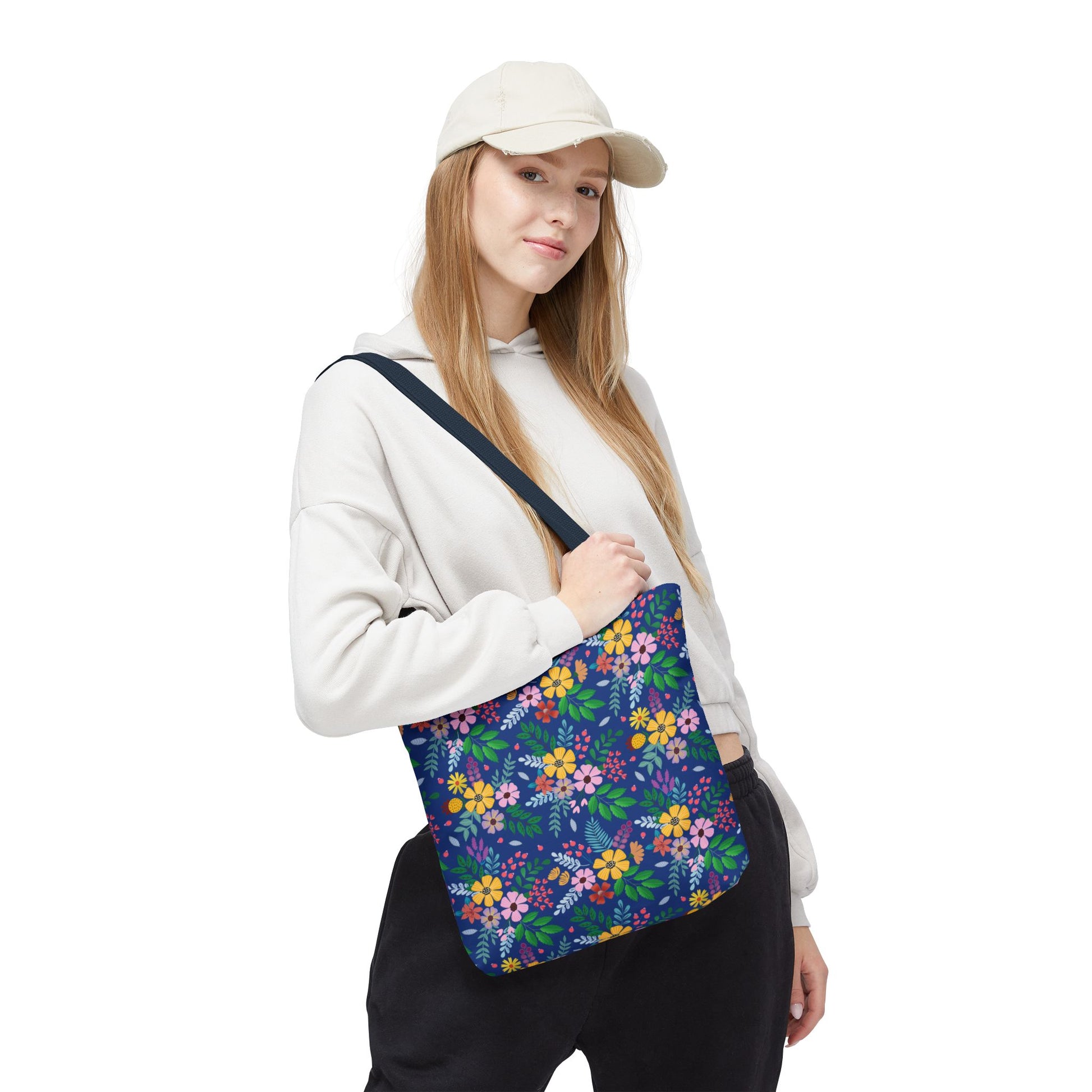 Young woman models the Floral Tote Bag with a bright, floral print on a dark navy background, complementing her casual light hoodie and dark pants. Ideal for adding a cheerful touch to summer outings.