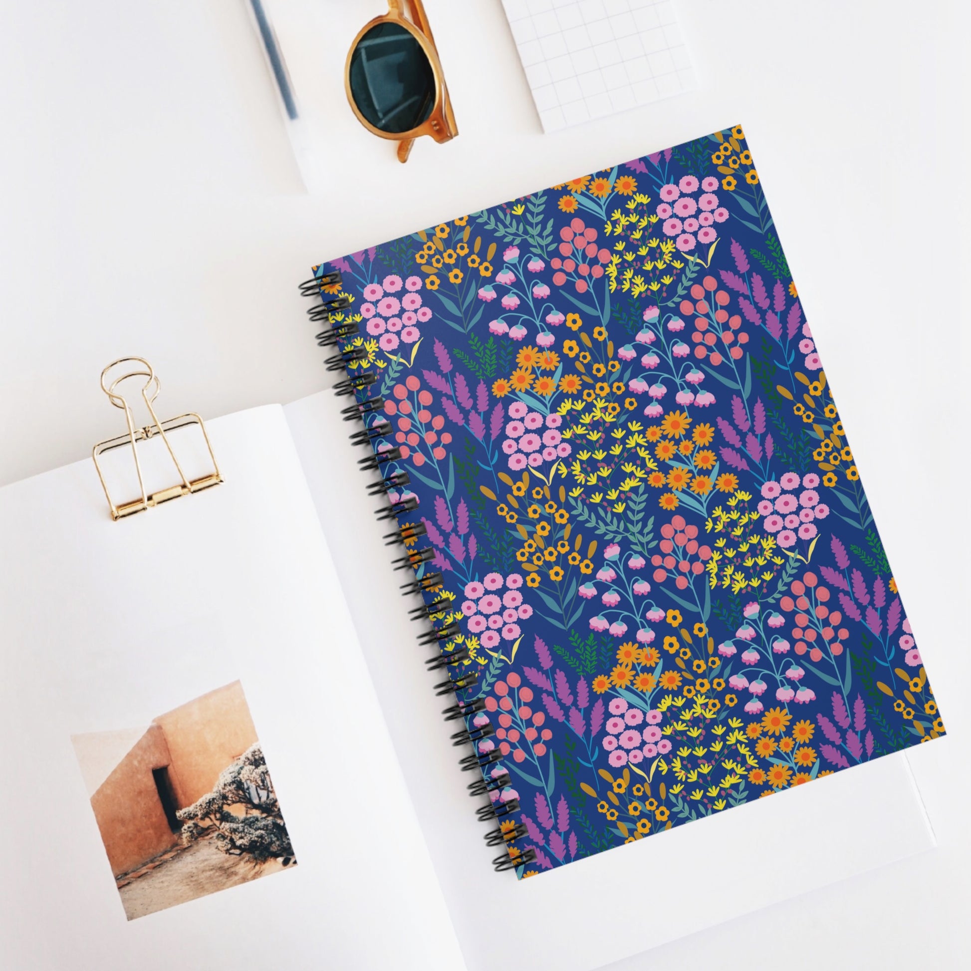 Floral Spiral Notebook with vibrant wildflower cover rests on an open magazine, accompanied by sunglasses and a notepad, embodying FireBrook Designs' joyful, stylish creativity for everyday inspiration.