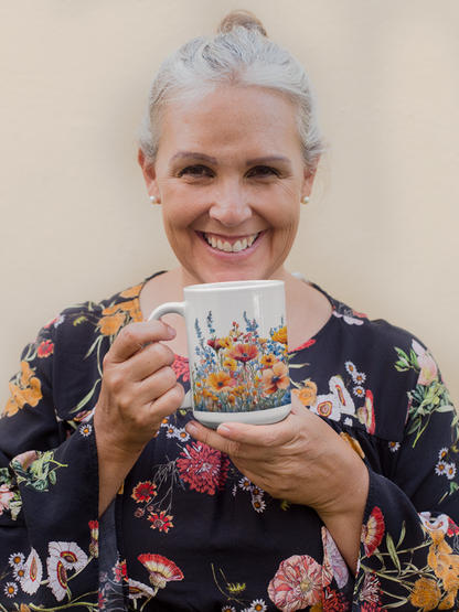 A woman smiles while holding the Wildflowers Mug (15oz), showcasing its vibrant floral design. This ceramic mug from FireBrook Designs offers a delightful, stylish touch to any beverage experience.